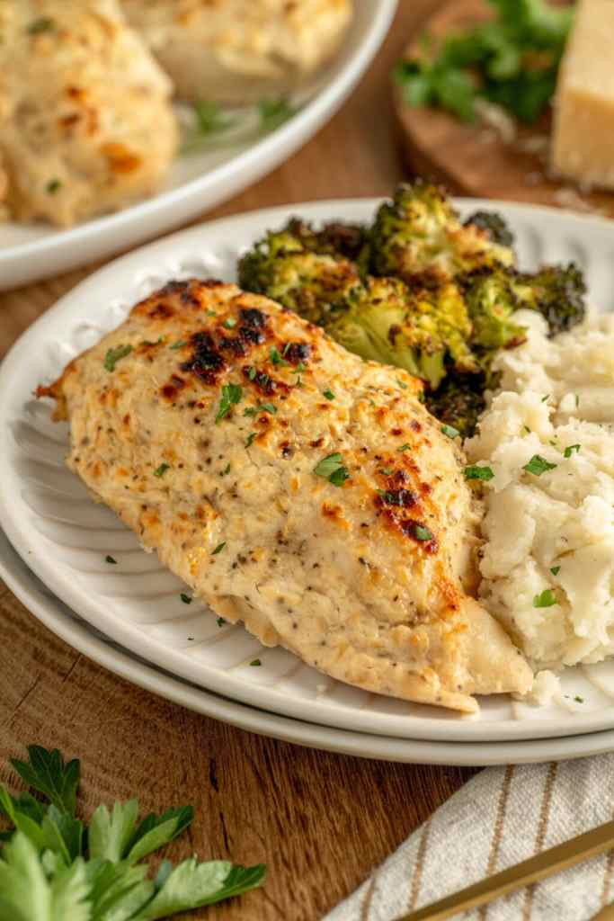 A plate of creamy garlic baked chicken breast served with steamed broccoli and mashed potatoes, garnished with fresh parsley.