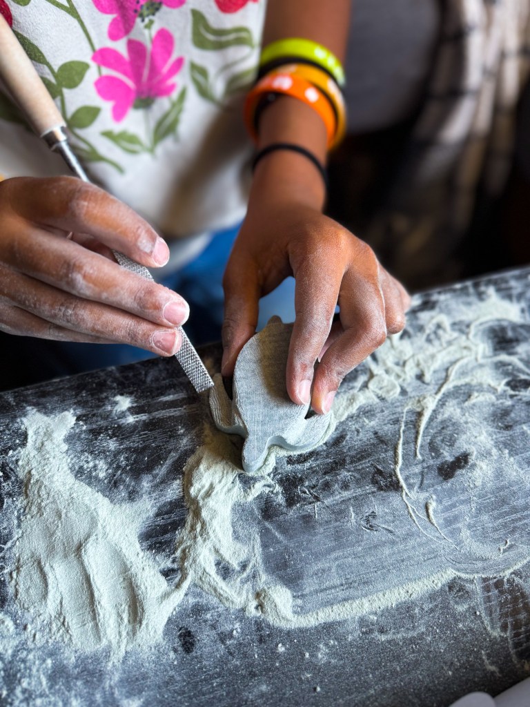 A close-up of a person's hands sculpting a piece of clay on a work surface covered in flour-like dust, using a carving tool.