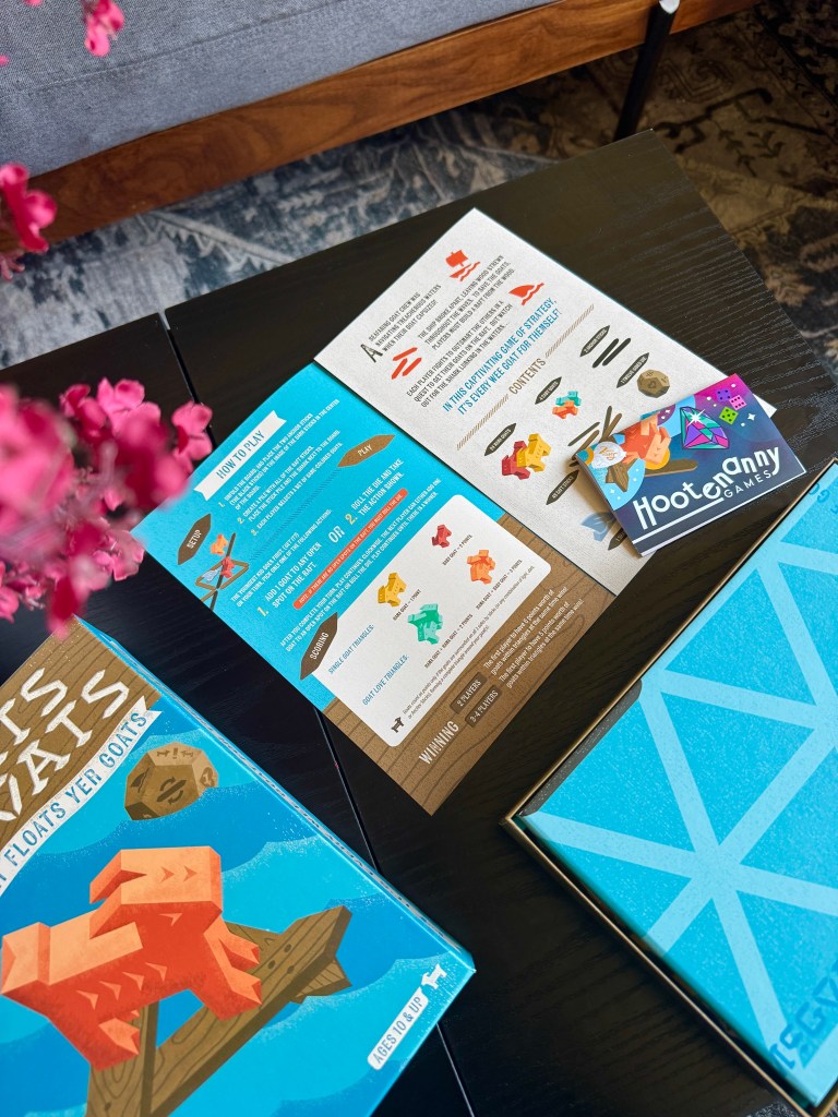 A tabletop game setup featuring a vibrant game box labeled 'It Floats Yer Goats', instruction sheets detailing how to play, and a small business card for Hootenanny Games, placed on a black table with a decorative floral arrangement in the foreground.