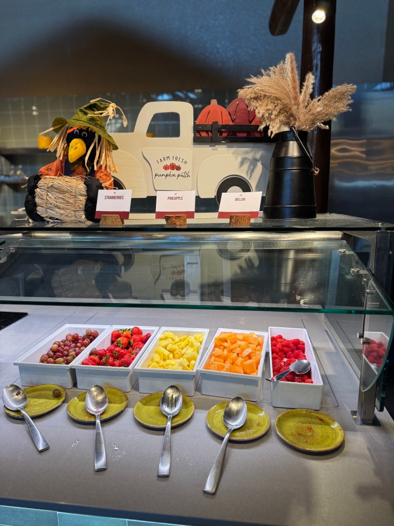 A colorful display of fresh fruits including strawberries, pineapple, cantaloupe, and melons, organized in small white containers on a buffet table, accompanied by decorative elements like a scarecrow and a wooden truck.