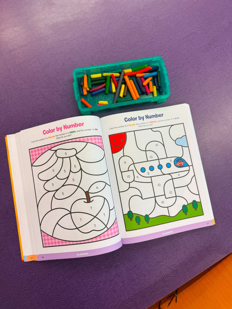 A color by number activity book opened to two pages with coloring prompts, accompanied by a box of crayons.