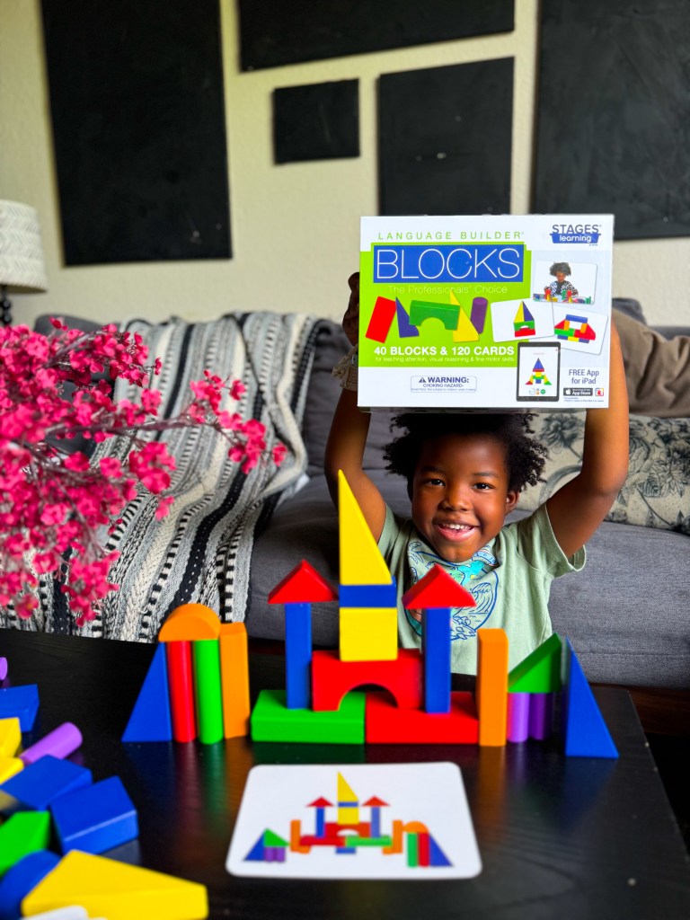 A joyful child holding a box of 'Language Builder Blocks' while playing with colorful building blocks on a table. A completed block structure is visible in front of the child, alongside an instruction card.