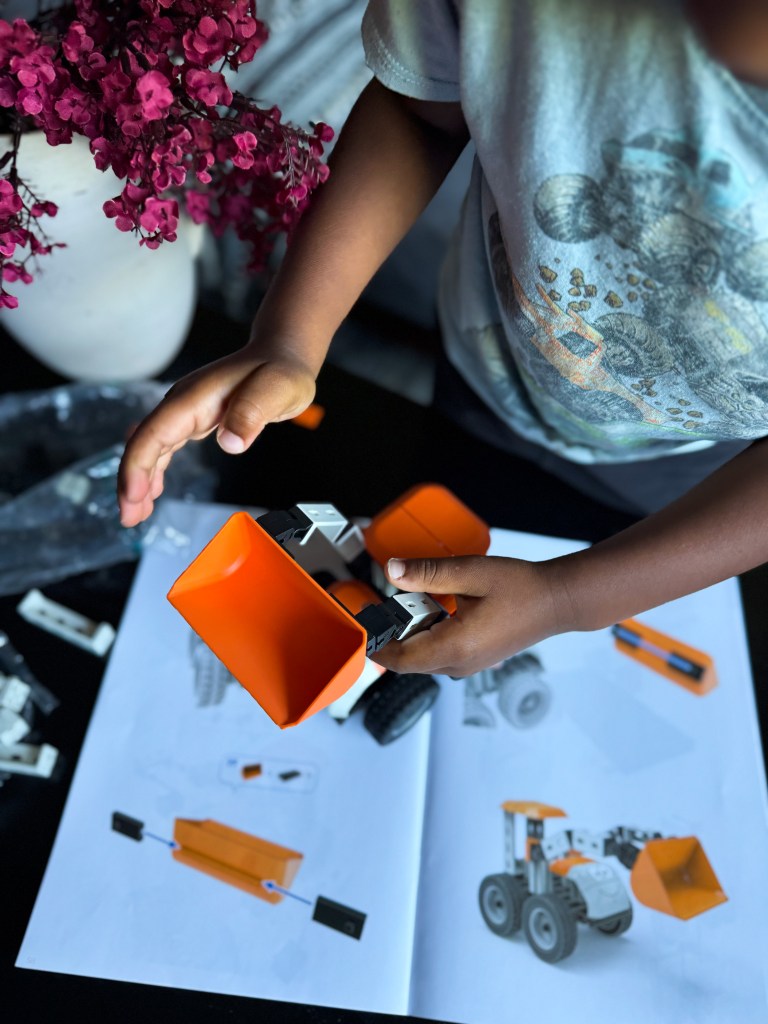 A child holding an orange construction vehicle toy with a detailed instruction manual on a table, alongside a pink flower arrangement.