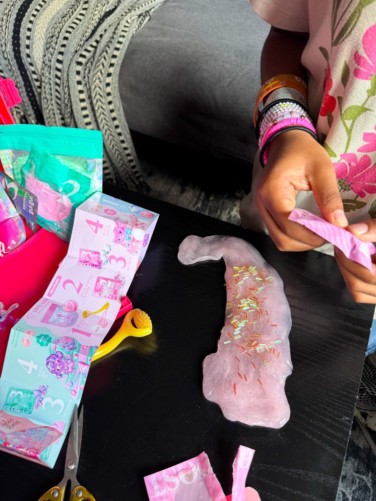 A child's hands working with pink slime on a table, surrounded by colorful toy packaging and accessories.