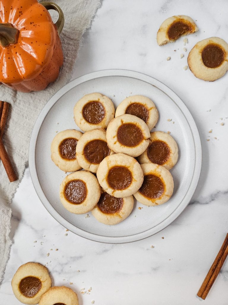 A plate of Pumpkin Pie Thumbprint Cookies with a cozy pumpkin-shaped mug and cinnamon sticks on a marble surface.
