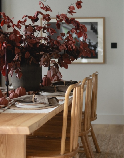 A cozy fall dining table set with a rustic wooden table, accented by a large vase of deep red leaves, small pumpkins, and neutral tableware, creating a warm and inviting atmosphere.