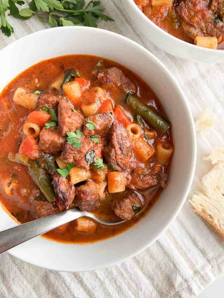 A bowl of hearty beef soup featuring tender chunks of beef, pasta, carrots, and green beans in a rich tomato broth, garnished with fresh herbs.
