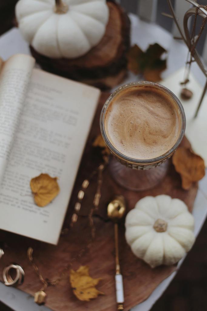 A cozy fall-themed flat lay featuring a glass of coffee, a partially open book, white pumpkins, golden leaves, and a decorative spoon, all arranged on a wooden surface.