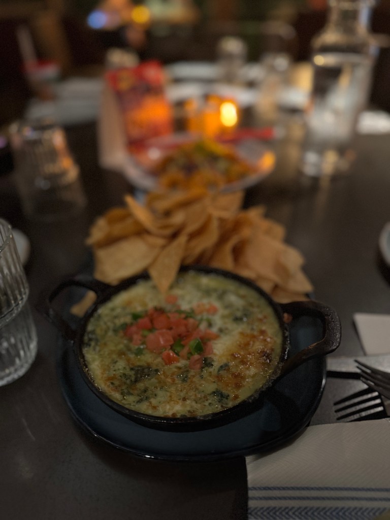 A skillet of baked spinach dip topped with diced tomatoes and scallions, served with tortilla chips on a dark table setting. Fireside Restaurant at Great Wolf Lodge in Grapevine, TX. 