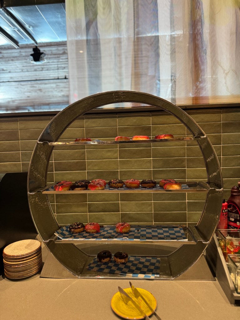 A tiered display of colorful donuts at the Great Wolf Lodge in Grapevine, TX dining area, with pink and chocolate frosted varieties, set against a green tiled background.