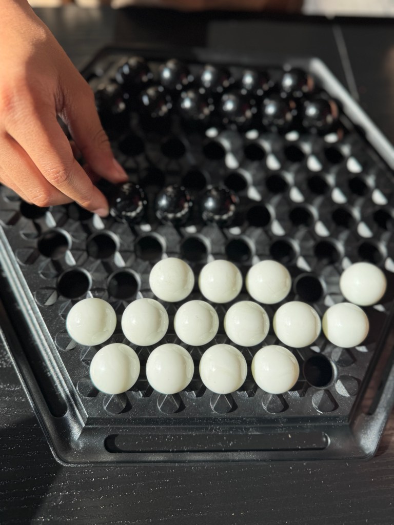 A close-up of a hand moving a black marble on a hexagonal board game, featuring white marbles arranged in a grid pattern.
