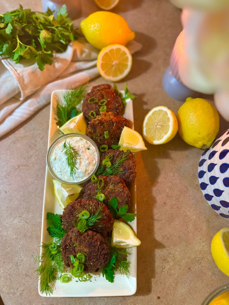 A platter of freshly fried salmon patties garnished with green onions and fresh herbs, accompanied by lemon wedges and a small bowl of creamy dip.