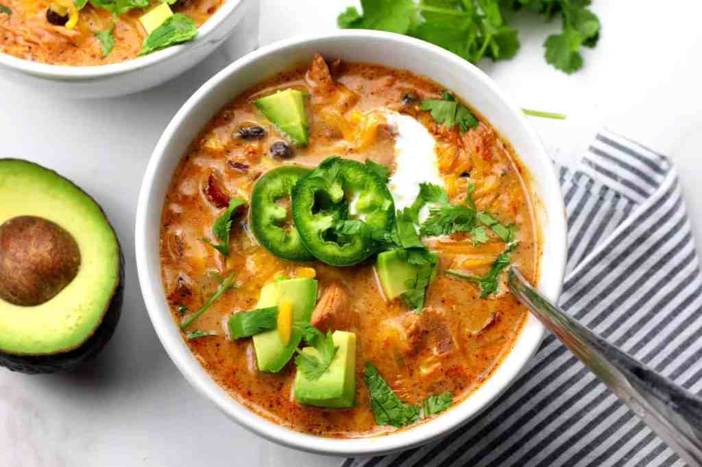 A bowl of creamy chicken soup topped with fresh cilantro, avocado, and slices of jalapeño, served with an avocado half and a striped napkin in the background.