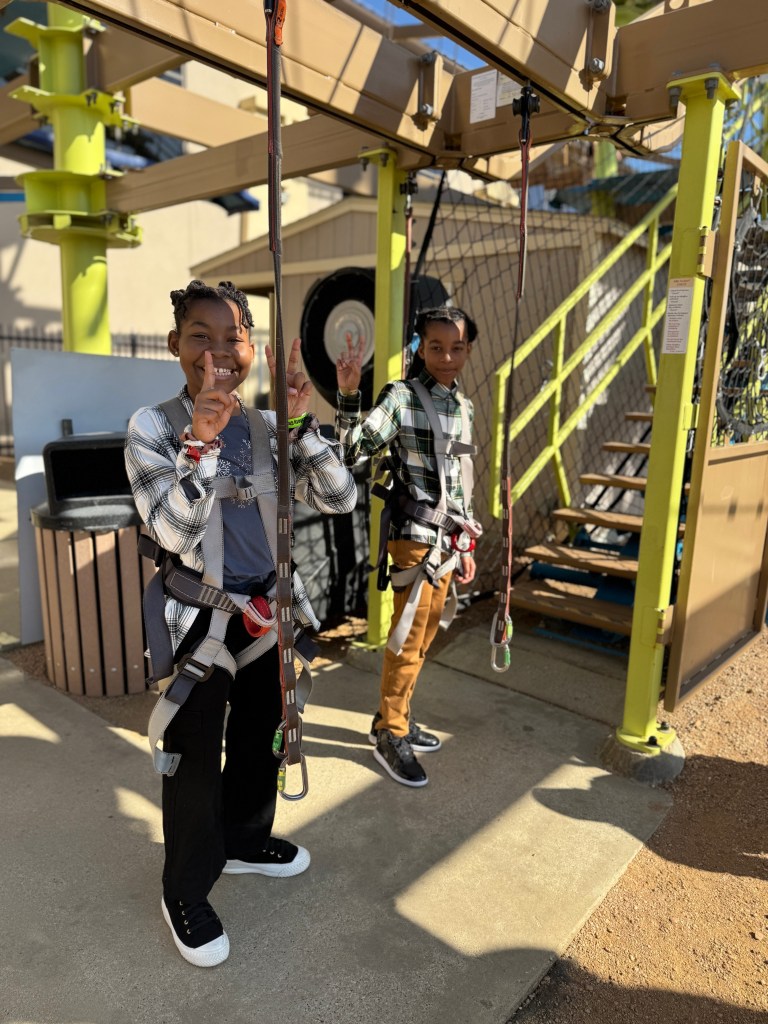Two children smiling and posing with harnesses before an outdoor adventure course, standing on a concrete surface near a safety net and climbing structure.