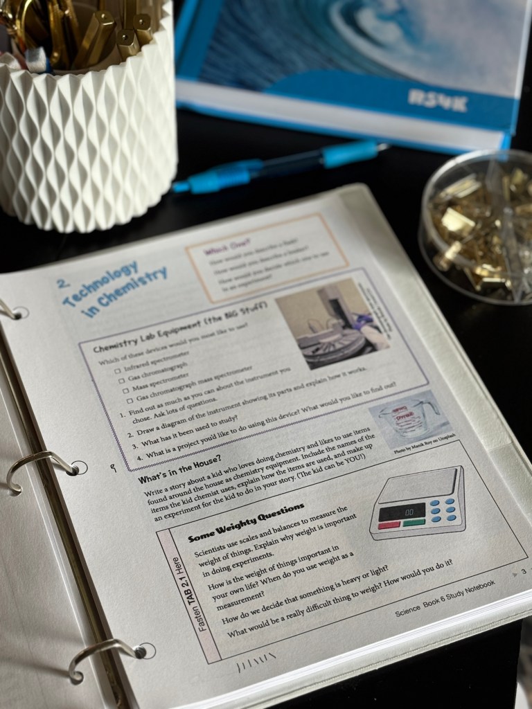 A close-up of an open page from the Real Science 4 Kids Book 6 Study Notebook featuring a section titled 'Technology in Chemistry,' with questions and prompts for students about chemistry lab equipment and experimental criteria. A pen and a container of paper clips are also visible in the background.