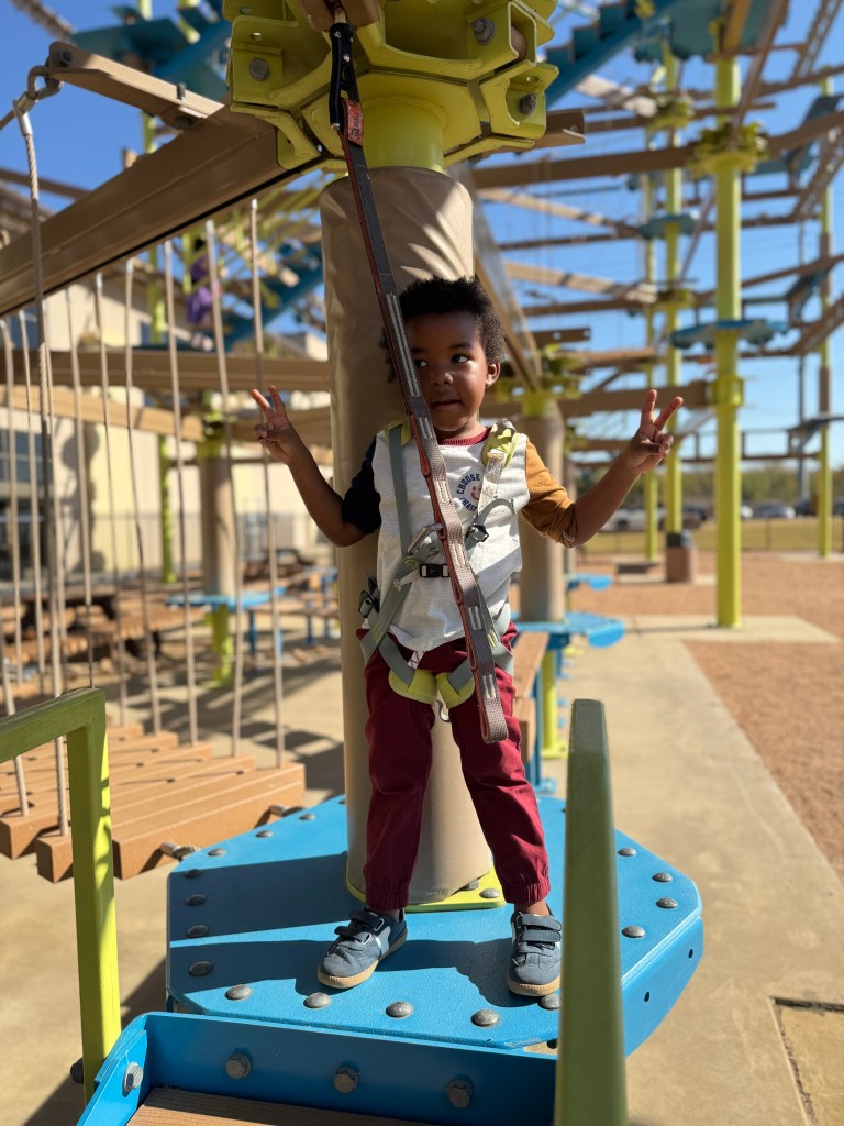 A young child wearing a harness stands on a platform in an adventure ropes course, smiling and making a peace sign with one hand.