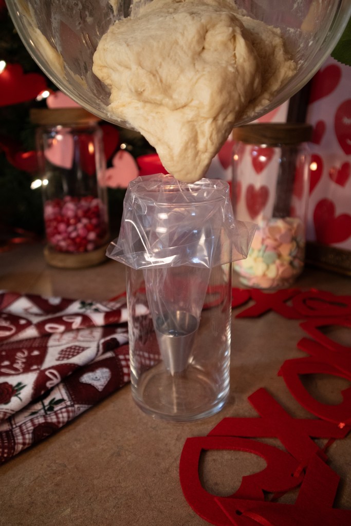 Dough being poured from a mixing bowl into a piping bag positioned in a glass, with colorful jars and decorative items in the background.