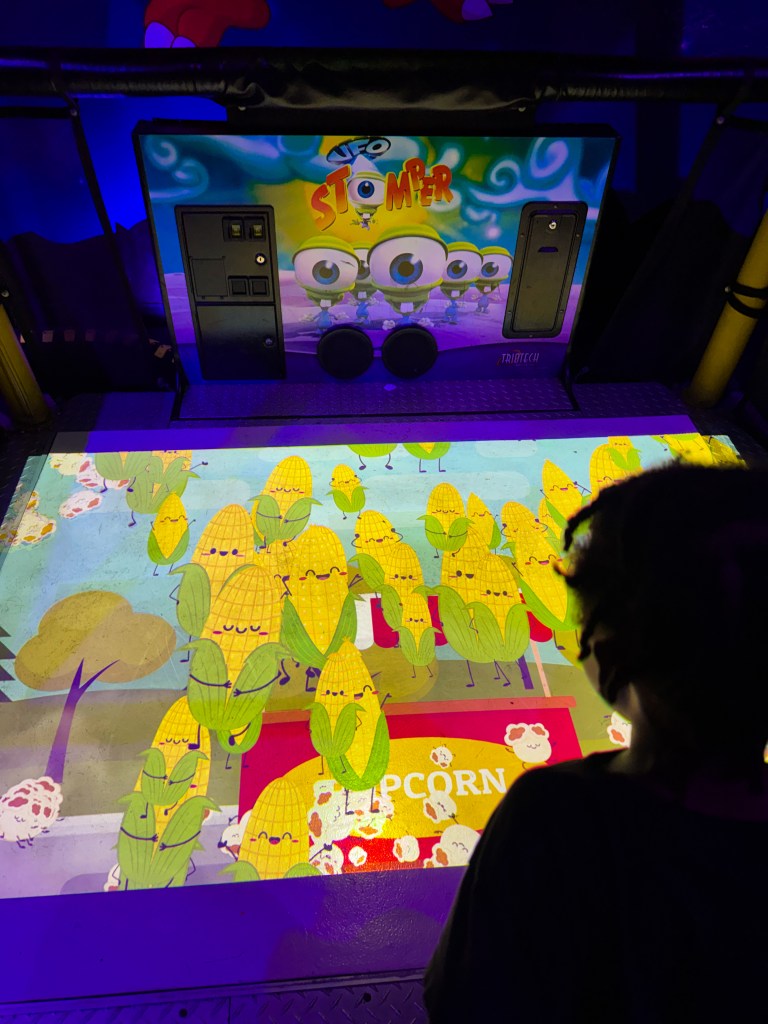 A back view of a child interacting with a colorful interactive game called 'UFO Stomper,' featuring animated corn characters and popcorn graphics on the screen.