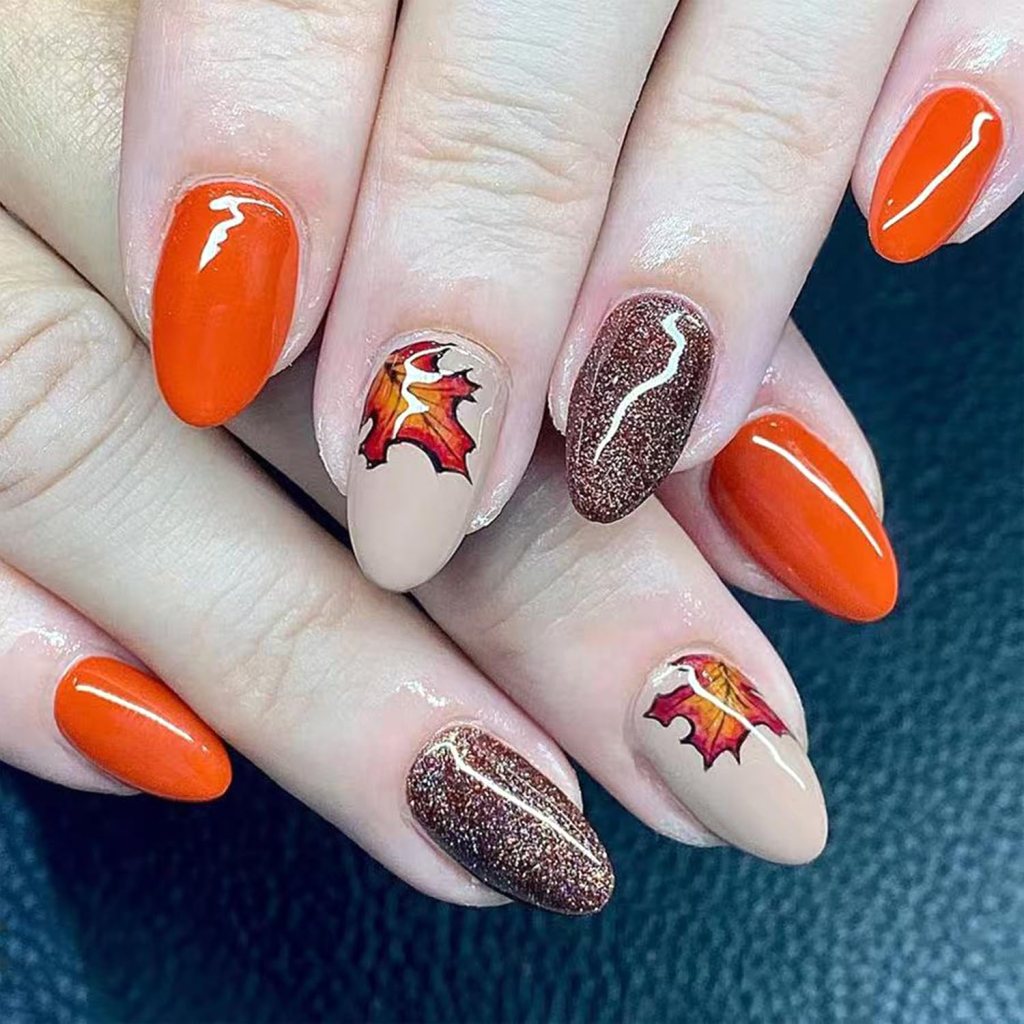 Close-up of festive Thanksgiving nails featuring rich orange and nude shades, with intricate fall leaf designs and a shimmery brown accent nail.