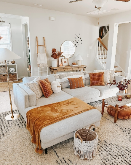 A cozy living room featuring a comfortable sectional sofa adorned with warm, earthy-toned pillows and a plush throw blanket. A wooden coffee table displays seasonal decor, including a small arrangement of autumn foliage. The space is bright and inviting, with light-colored walls and a stylish rug.