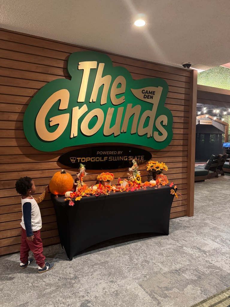 A cozy indoor space featuring 'The Grounds' game den sign, decorated with autumn-themed accents, including pumpkins and flowers. A child stands nearby, inspecting the festive display.
