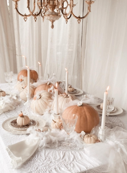 Elegant fall table setting featuring soft peach and white pumpkins, tall white candles, and crystal glassware on a delicate lace tablecloth.