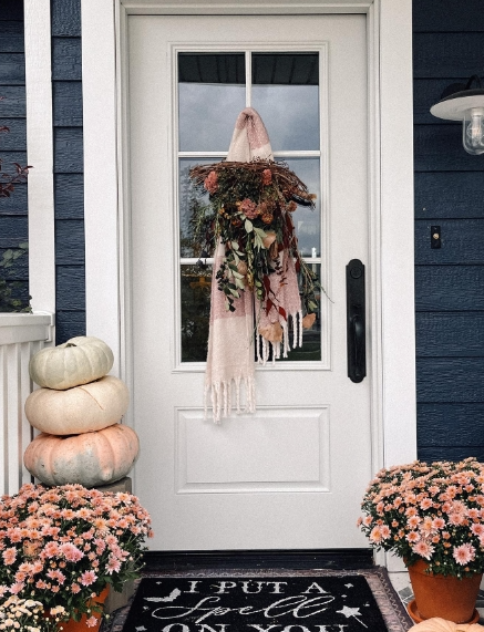 A cozy front porch featuring a white door with a floral wreath, pastel pumpkins stacked beside it, and vibrant pink mums on either side.