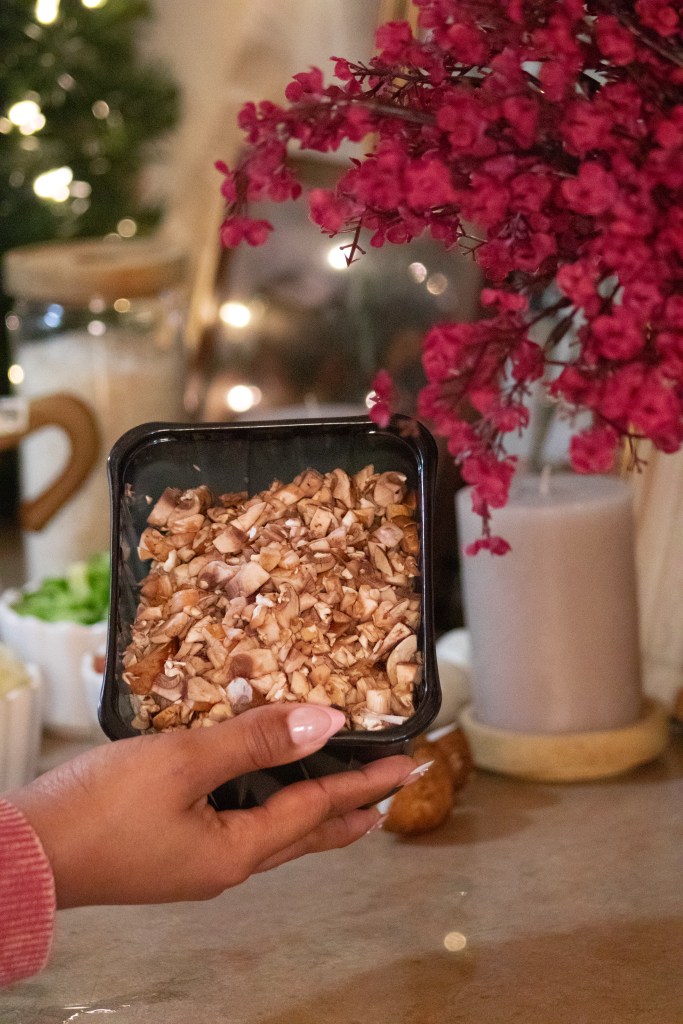 A hand holding a container of chopped mushrooms, with a blurred background featuring flowers and soft lighting.