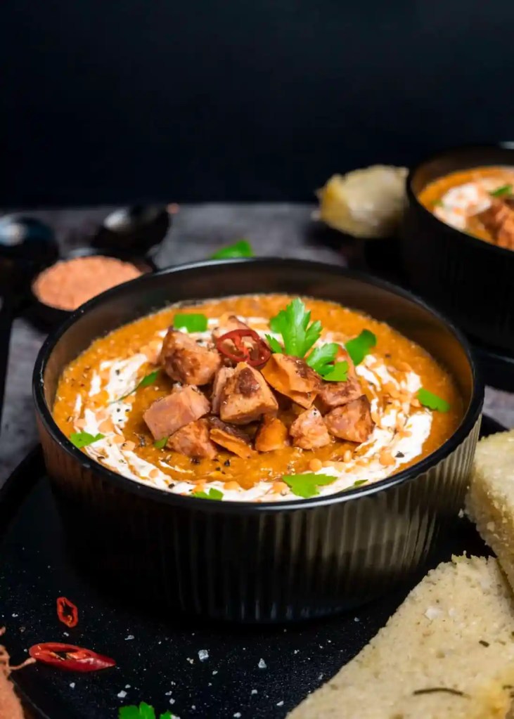 A bowl of creamy chickpea curry soup garnished with herbs and spices, accompanied by slices of bread.