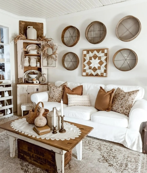 A cozy living room featuring a white sofa adorned with decorative cushions, a rustic coffee table with a lace doily, candles, and a woven basket. A wooden shelf in the background displays various decor items, while patterned wall art and circular wooden decor pieces enhance the warm atmosphere.