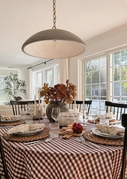 Thanksgiving tablescape featuring a checkered brown tablecloth, layered place settings with woven chargers, candles, and a centerpiece of fall foliage and decorative elements.