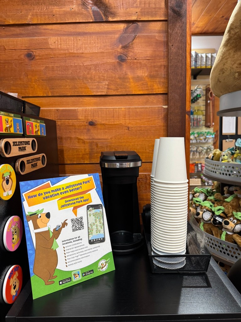 A coffee machine next to a stack of paper cups on a black table, with a promotional sign for Jellystone Park highlighting the app download and vacation information.