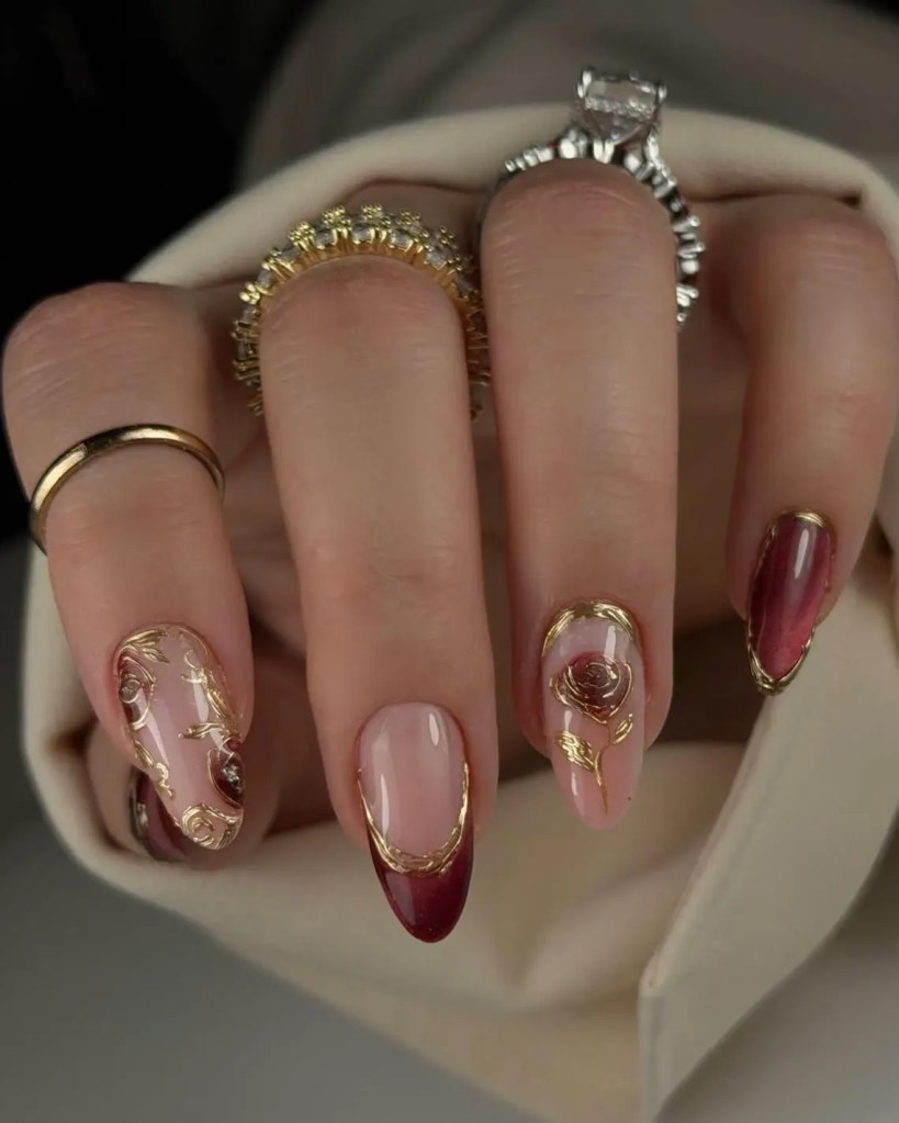 A close-up of a hand with elegantly manicured nails featuring a combination of nude and deep red colors, along with gold embellishments, and multiple decorative rings.