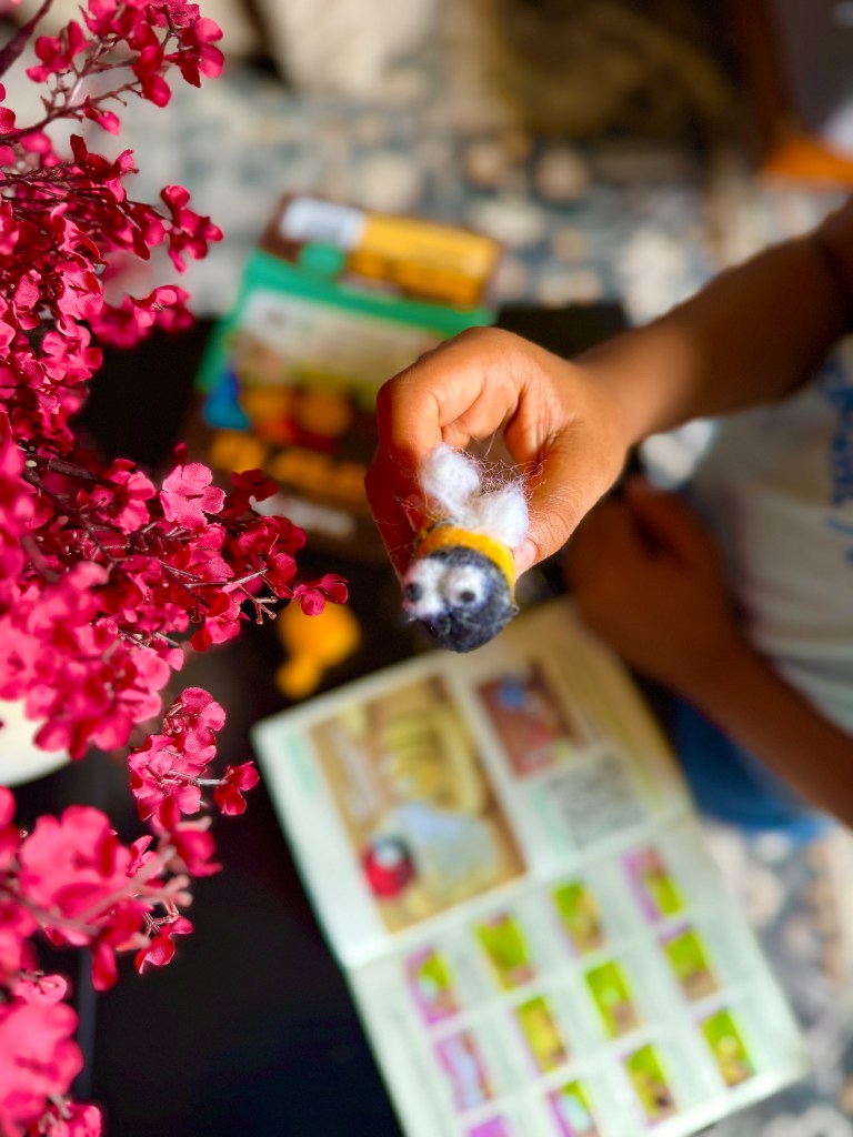 A child's hand holding a felted bee figure, with a colorful flower arrangement and a needle felting instruction booklet in the background.