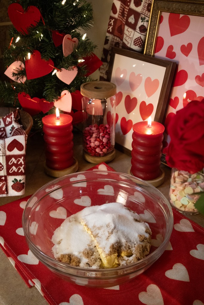 A glass bowl with butter and sugars, surrounded by a romantic Valentine's Day setting featuring a small Christmas tree adorned with hearts, red candles, and heart-themed decorations.