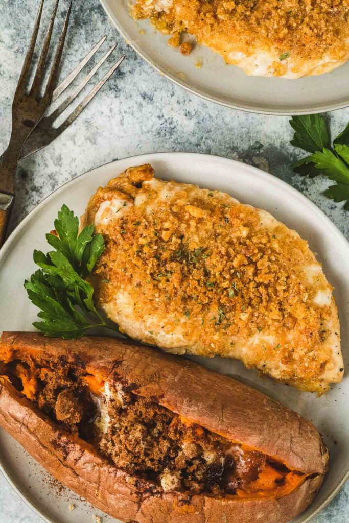 A plate featuring crispy baked chicken breast topped with a golden brown crumb coating, alongside a baked sweet potato filled with brown sugar and marshmallow topping, garnished with fresh parsley.