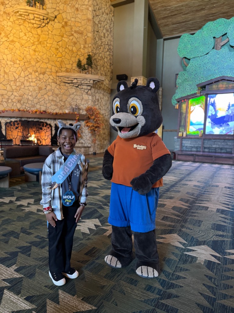A child standing next to Brinley Bear, a character mascot at Great Wolf Lodge, in a cozy lodge lobby decorated with a stone fireplace and autumn-themed decor. The child is wearing a sash and has a joyful expression.