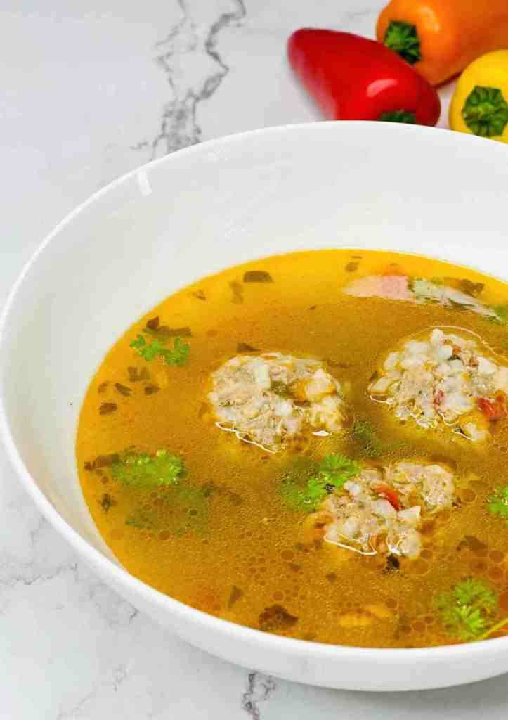 A bowl of clear soup with meatballs and herbs, garnished with fresh cilantro, on a marble surface, accompanied by colorful bell peppers.