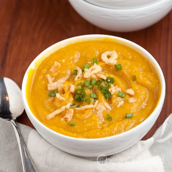 Bowl of creamy butternut squash soup topped with crispy fried onions and green onions, placed on a wooden table with a spoon and cloth napkin.