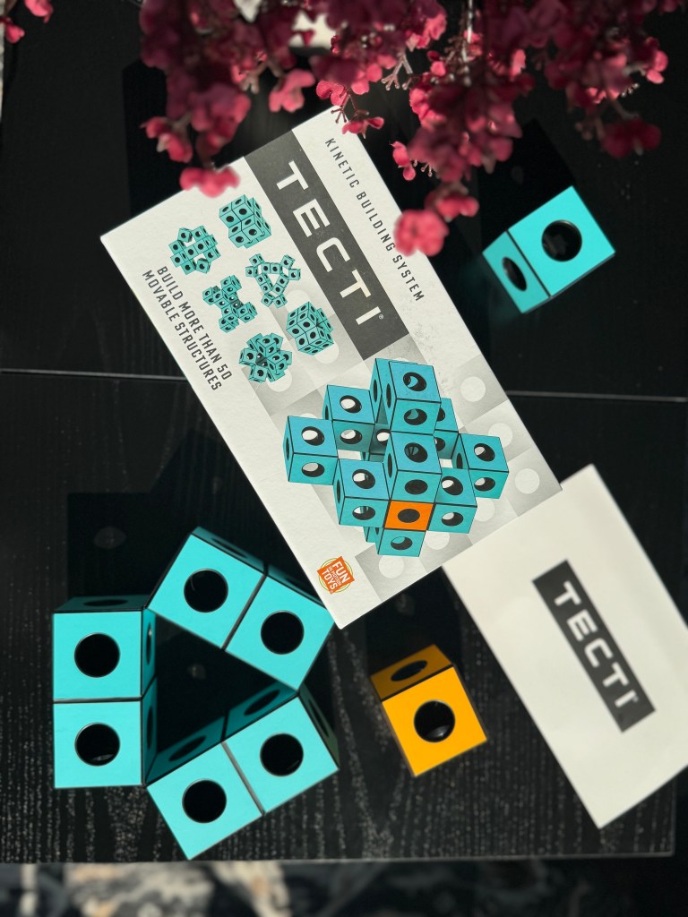 A colorful display of the Tecti kinetic building system, featuring turquoise and black cubes with circular openings, alongside the product box on a black table.