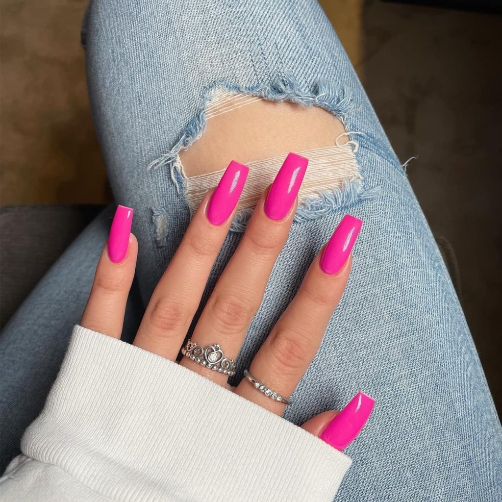 A close-up view of a hand with long, bright pink nails resting on a knee. The person is wearing a light grey sweater, and the knee is covered with distressed denim jeans featuring a large rip.