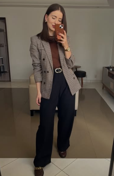 A young woman poses in a stylish outfit featuring a checked blazer over a brown top, paired with black wide-leg trousers and a matching belt. The setting is a modern, minimalistic room.
