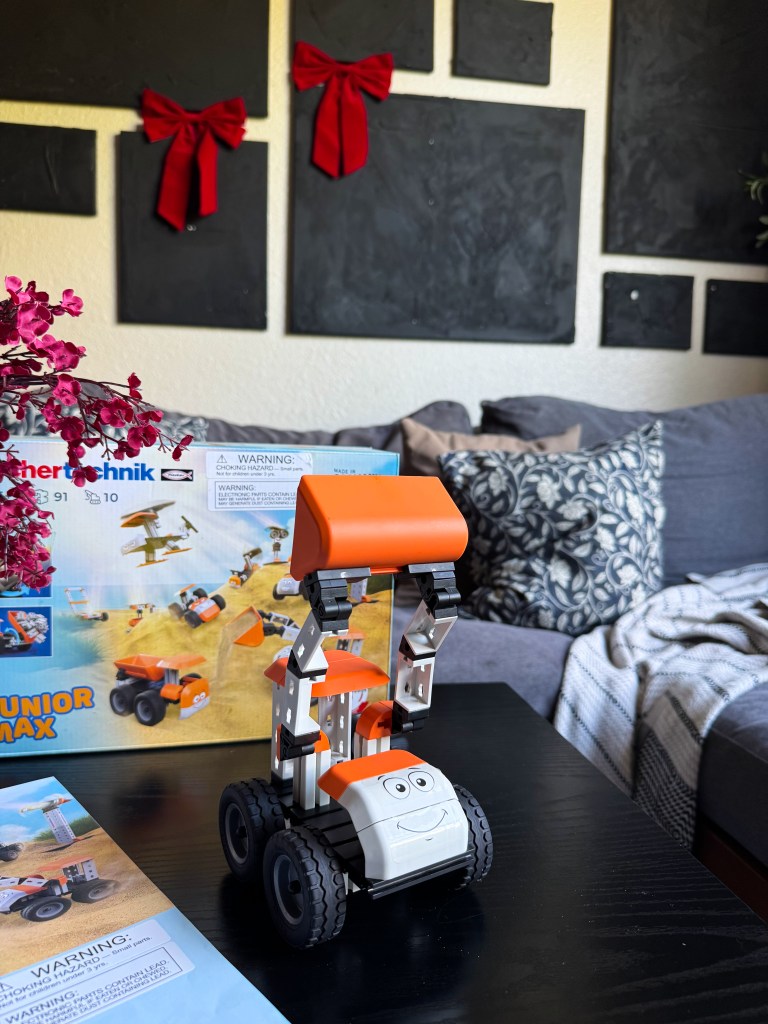 A colorful toy construction vehicle with a smiling face, displayed on a black table with a flower, a box, and a manual in the background. The backdrop features a couch and wall art with red bows.