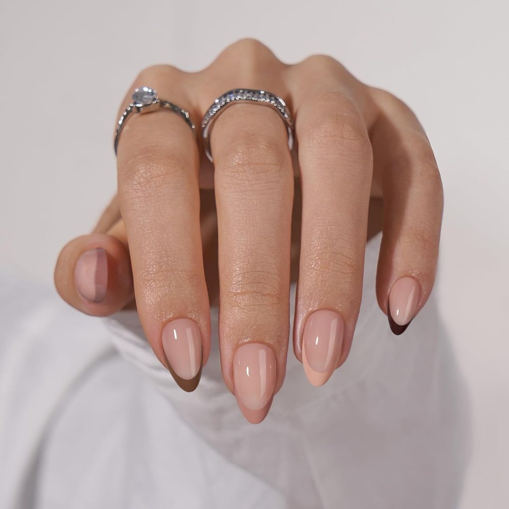 Close-up of a hand displaying stylish, nude-colored nails with a glossy finish, accessorized with two rings, one featuring a small gemstone.
