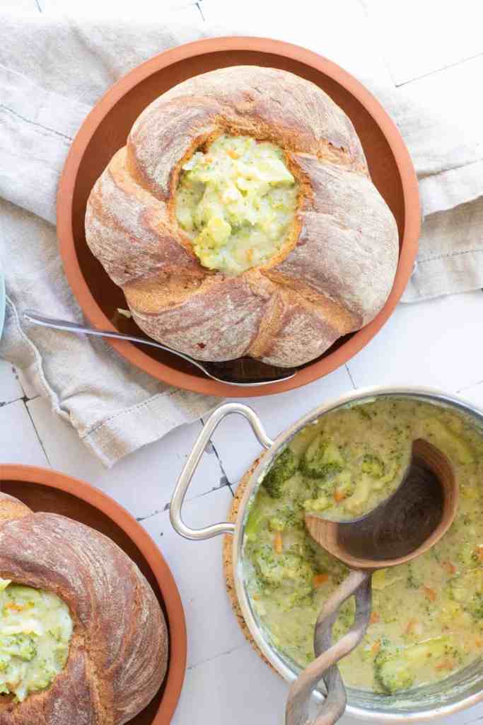A warm bowl of broccoli cheddar soup served in a sourdough bread bowl, with a pot of soup and a wooden spoon in the background.
