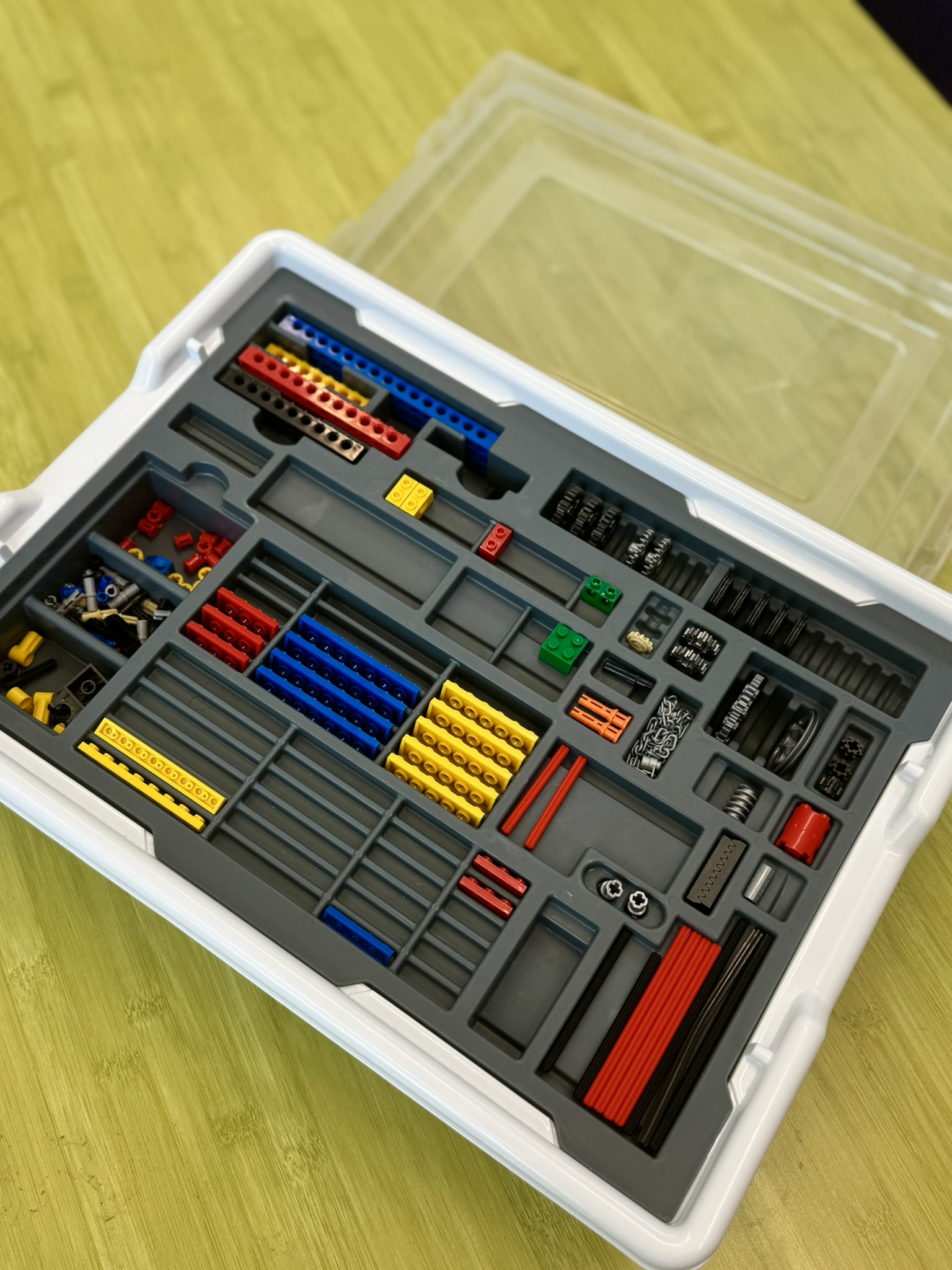 Plastic case with organized compartments containing various colored LEGO Technic pieces and gears on a wooden surface