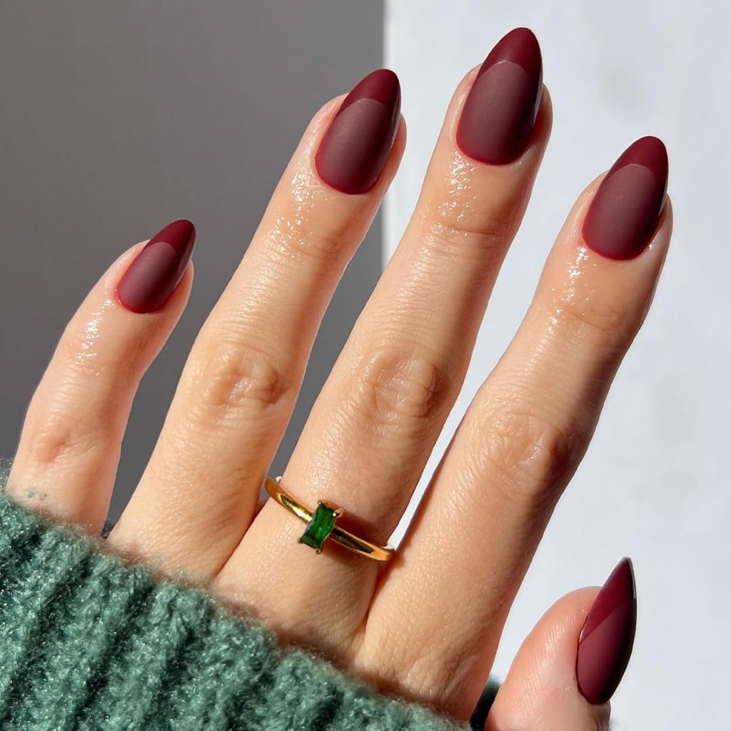 Close-up of a hand with well-manicured maroon matte nails featuring a contrasting glossy tip, wearing a gold ring with a green gemstone, partially covered by a green knitted sweater.