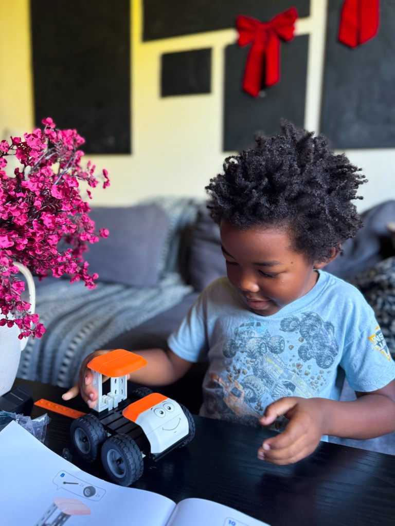 A young child playing with a toy truck on a table, surrounded by a pink flower arrangement and instructional booklet.