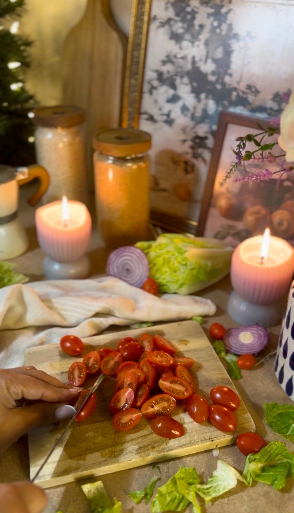 A person slicing cherry tomatoes on a wooden cutting board in a cozy kitchen setting, with candles, jars, and fresh vegetables in the background.