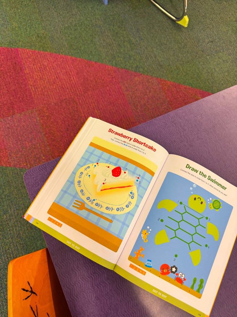 An open children's activity book displaying a page with a drawing of a strawberry shortcake on a plate and instructions for connecting dots to draw a swimmer turtle.